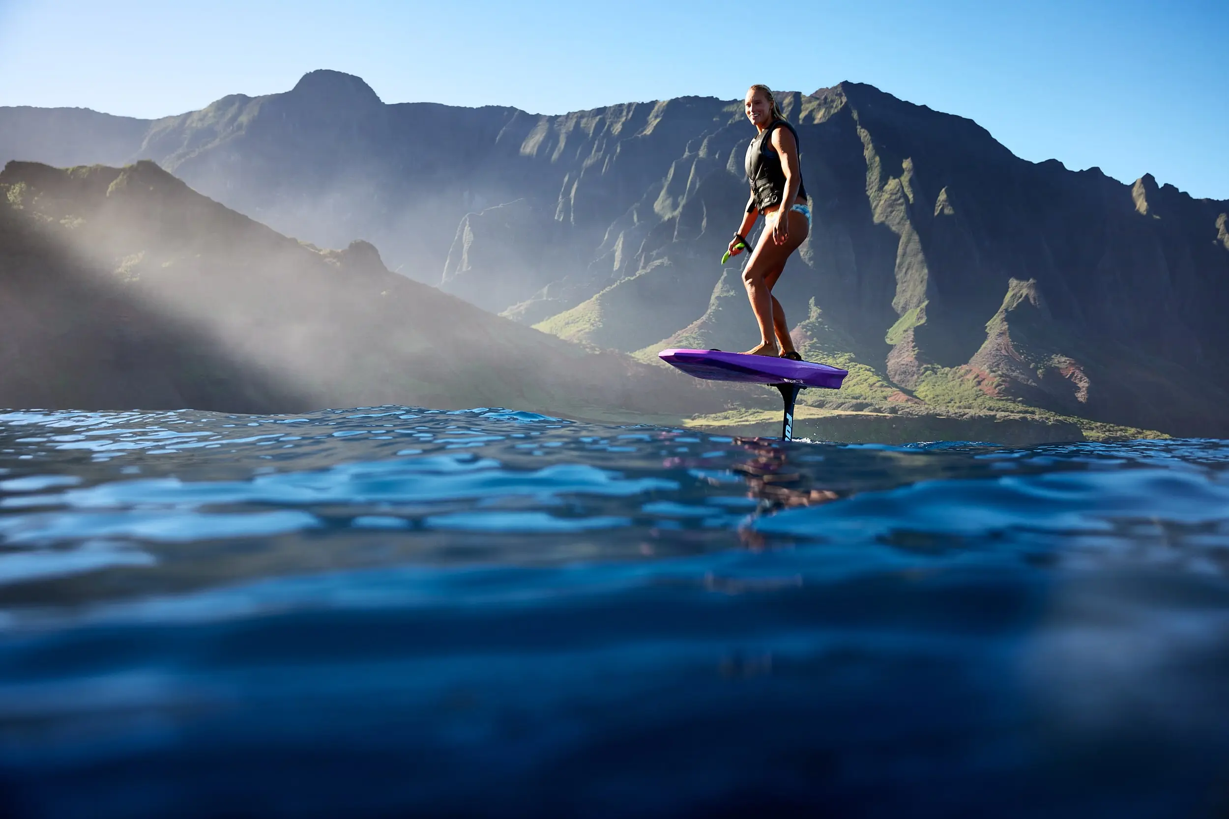 A profile shot of a female rider carving on the original FOIL board. The woman is smiling and moving through calm waters. Idyllic mountains are displayed in the background.