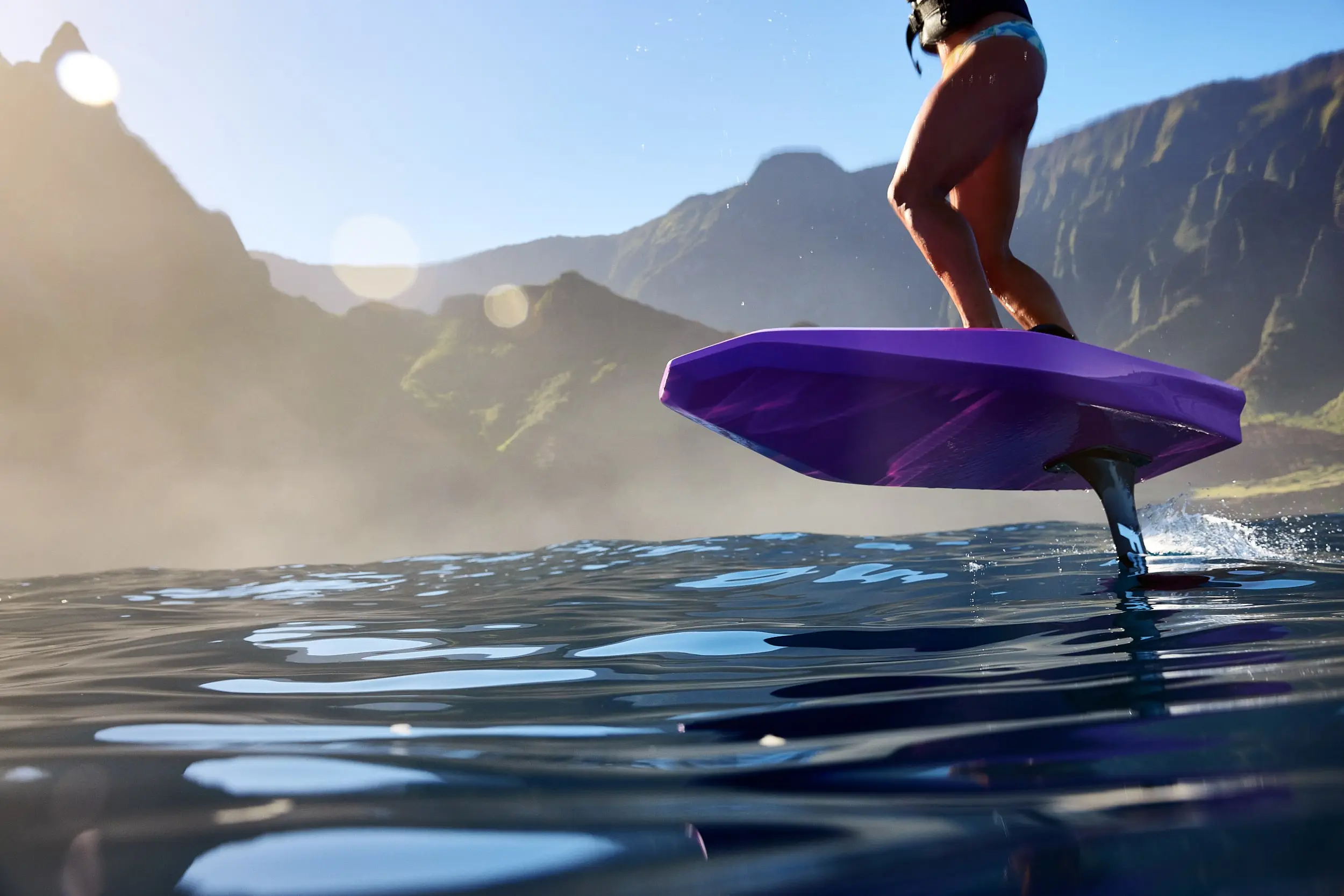 An image of a rider carving on the original FOIL model. The rider is carving in calm waters. The image is taken from the surface of the water, looking up at the rider. Mountains are pictured in the background.