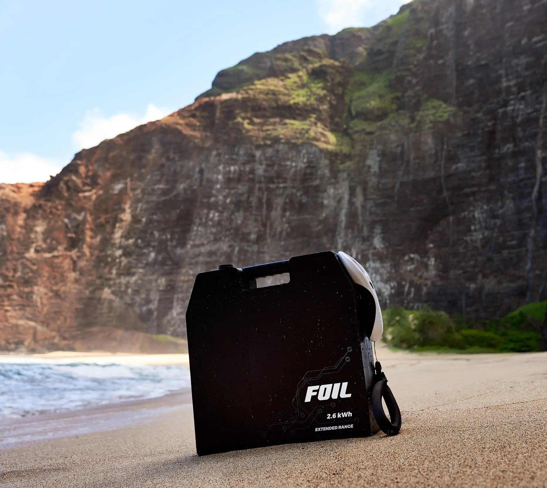 The FOIL 2.6 kWh battery resting on a beach. Breathtaking cliff walls, vibrant vegetation, and the ocean are visible in the background.