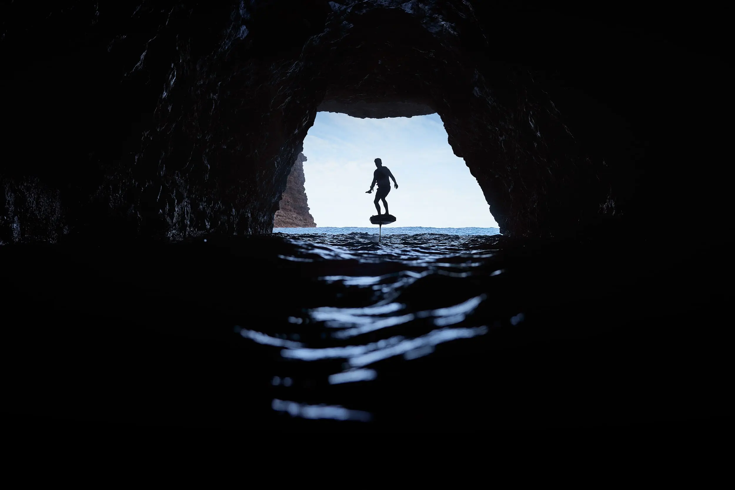 A FOIL rider cruising into a dark Hawaiian cave. The image is taken from inside the cave, and the ocean and horizon are visible in the background.