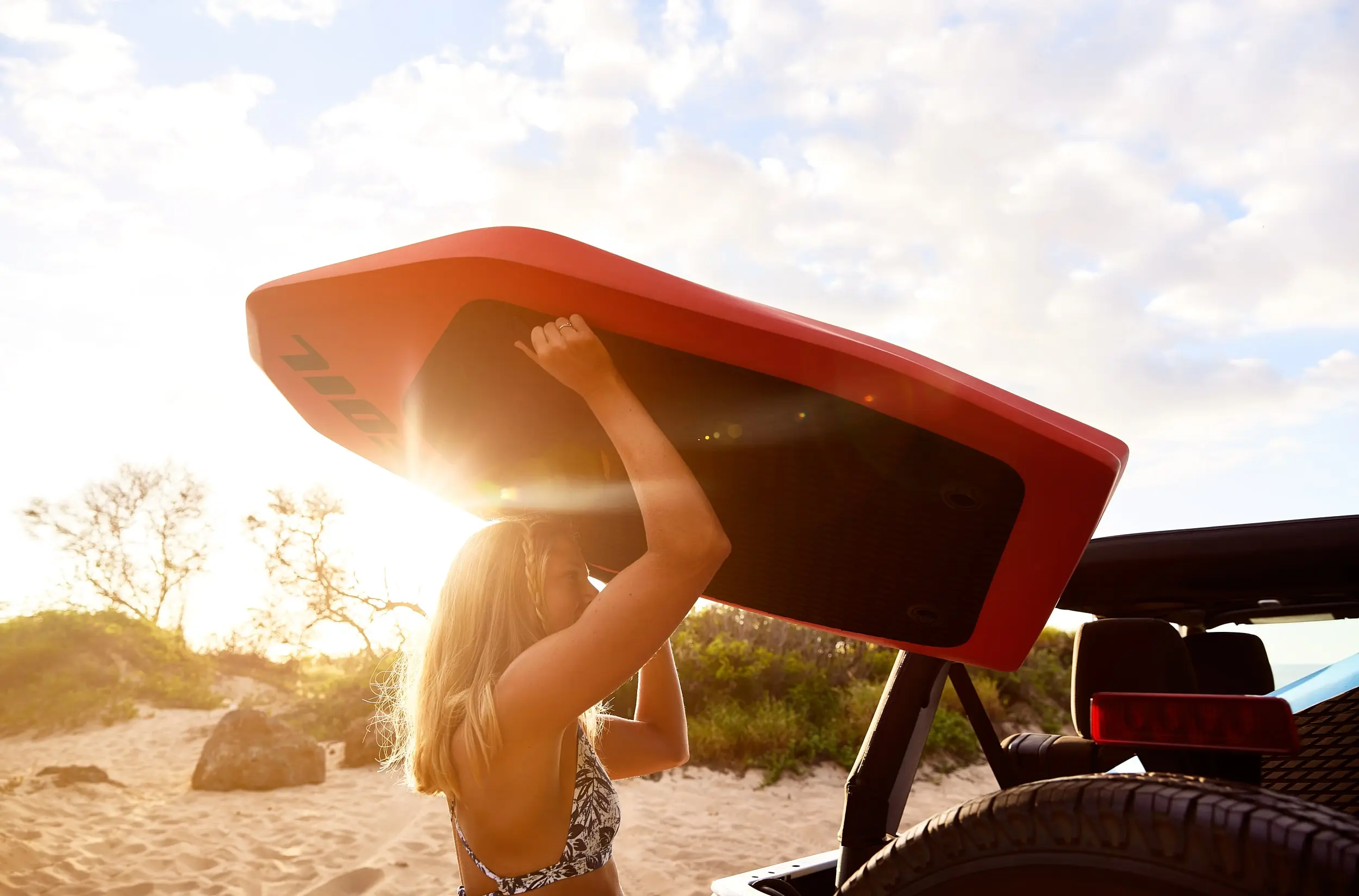 A woman lifting a FOIL board into the back of a jeep. Sunshine is piercing into the foreground from the background, and beach vegetation and sand are visible throughout the image.