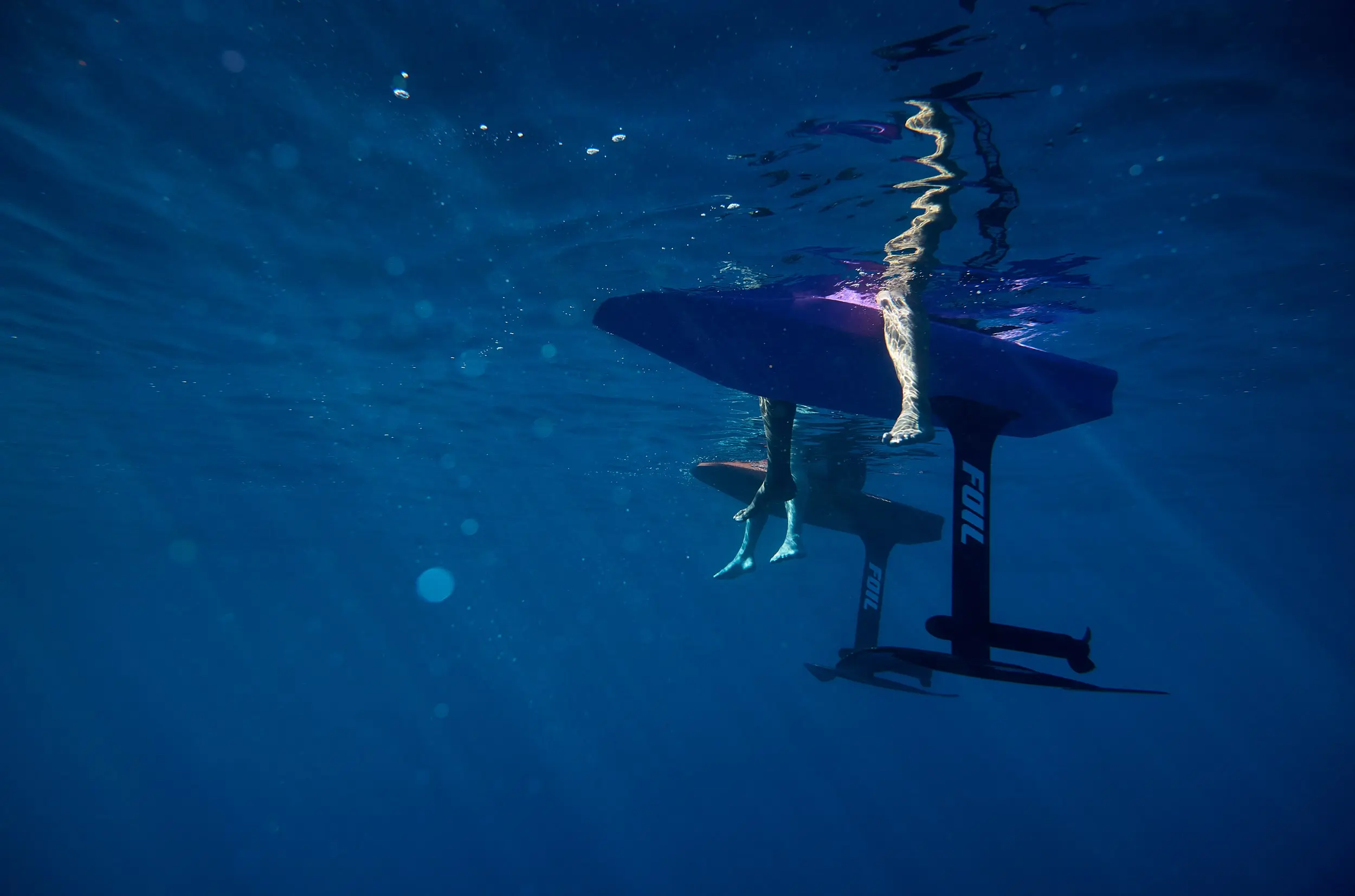 Underwater shot of two FOIL boards floating on the surface. The legs of two eFoil riders are positioned off the side of the board and sun rays are penetrating through the water’s surface.